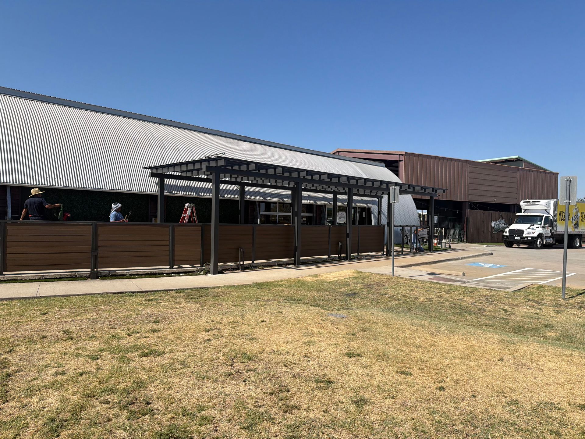 Outdoor market building with shaded patio, grassy foreground, and parked vehicles under a clear blue sky