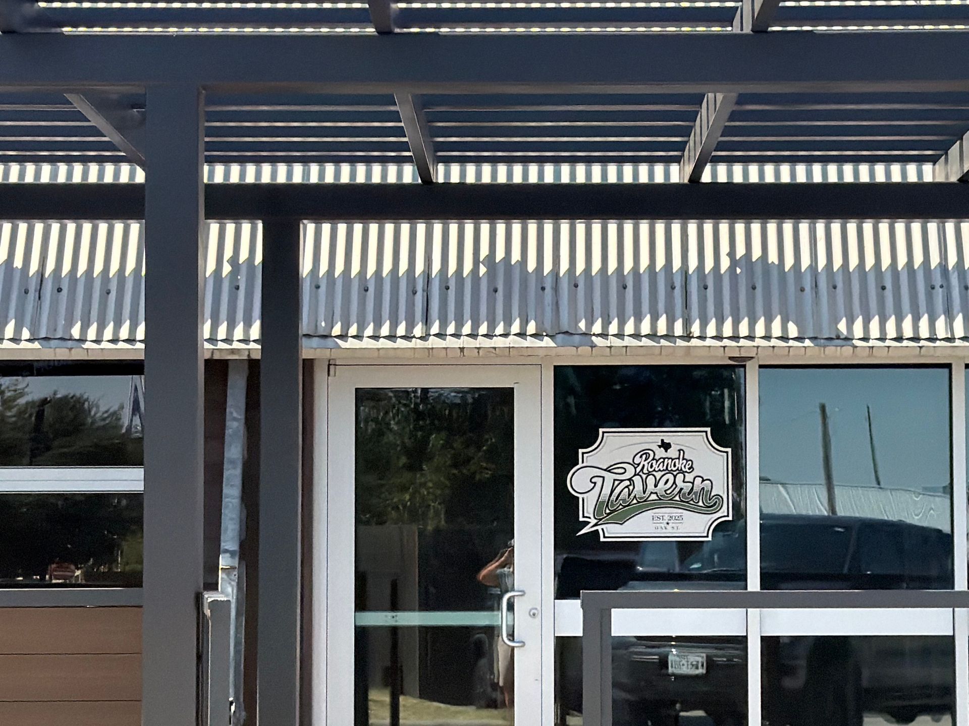 Click To See Roanoke Tavern's New Pergola Storefront entrance with blue awning, glass door, and window sign behind a metal railing at Roanoke Tavern, in Roanoke, Texas