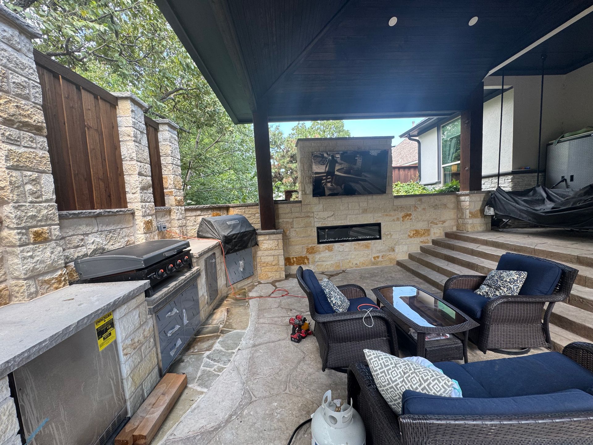 Covered outdoor patio with sectional seating, stone fireplace, grill, and stairs leading to a house.