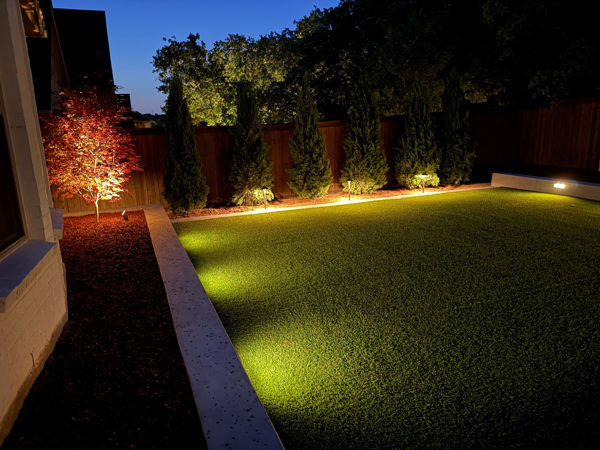 Illuminated backyard lawn at night with warm landscape lighting and a lit fence line