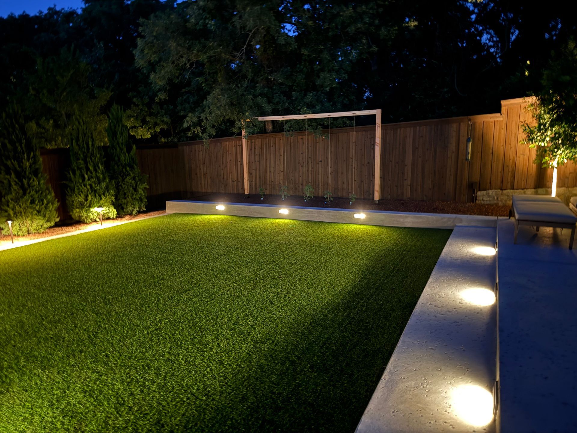 Lit backyard patio with green lawn, wooden fence, and ground lights at night