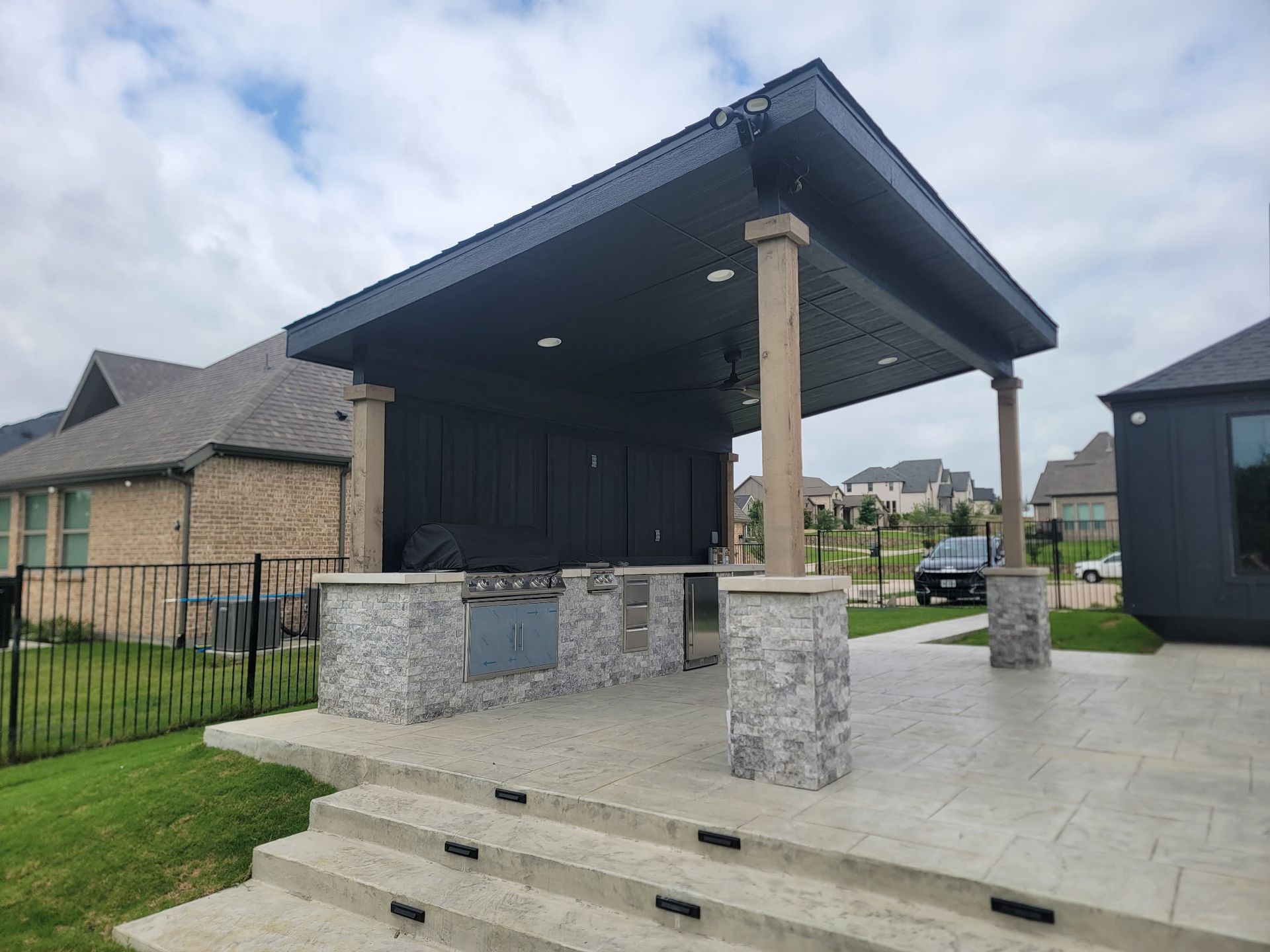 Covered outdoor patio with stone columns, tiled floor, and steps in a suburban neighborhood.