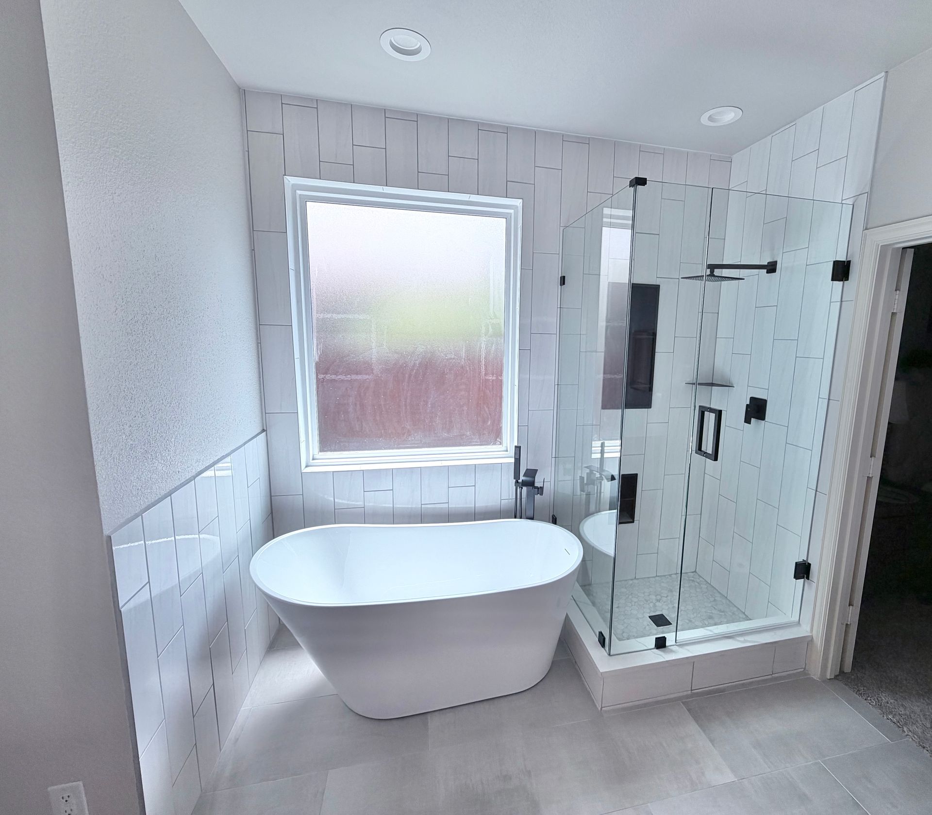 Bright bathroom with freestanding tub, glass shower, frosted window, and light gray tile.