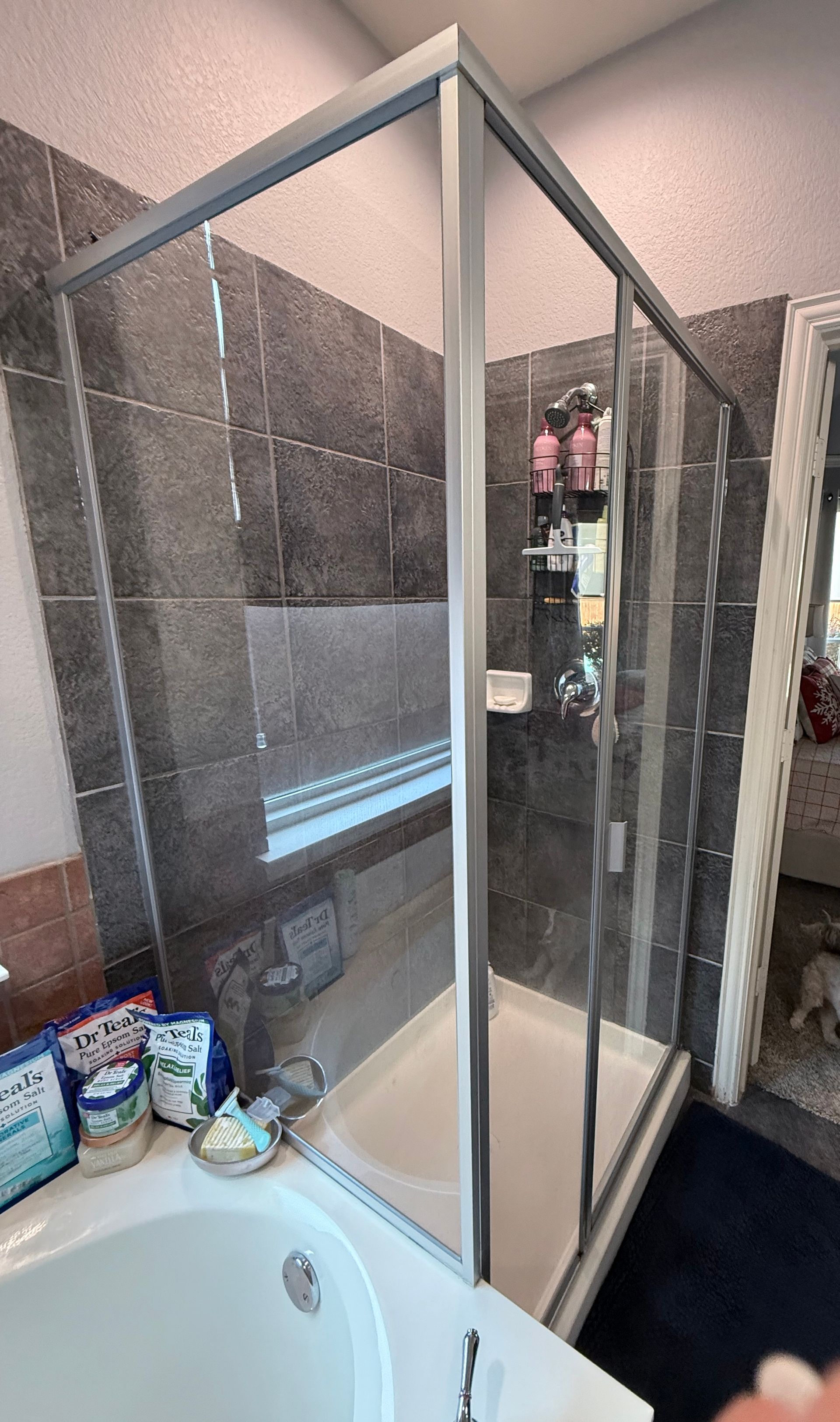 Glass shower enclosure in a bathroom beside a bathtub, with tiled walls and shelves of toiletries nearby