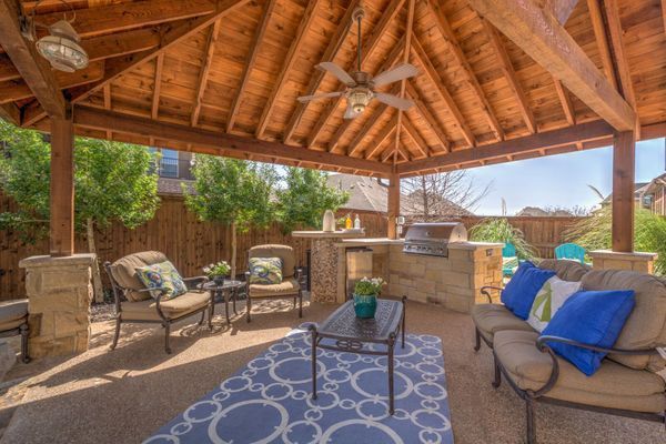 Covered backyard patio with wood-beam roof, seating, fire pit, and colorful cushions under a ceiling fan.