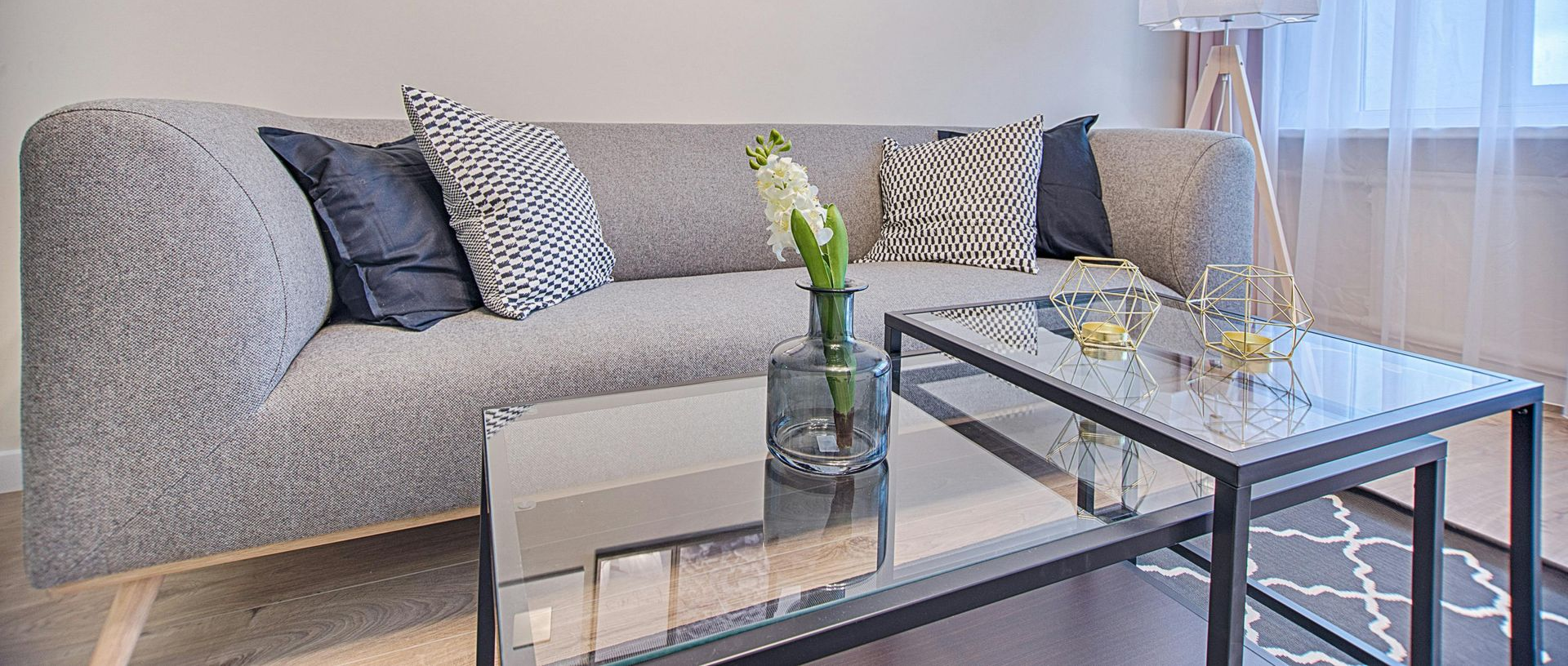 A grey fabric sofa with patterned cushions sits behind a tiered glass coffee table holding a vase with white flowers.