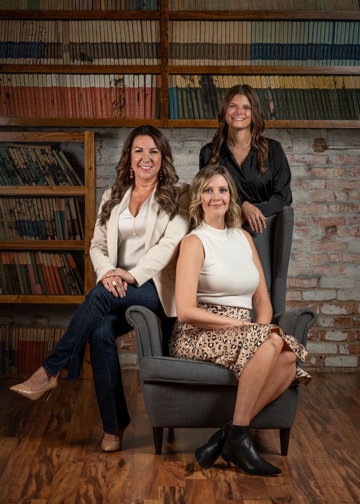 Three women posing in a rustic library, two seated and one standing behind, smiling at the camera