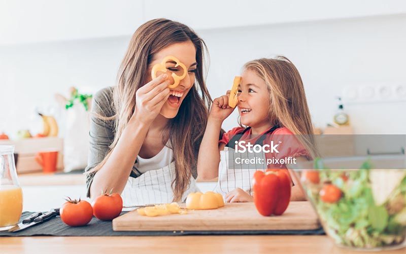 Woman and child making playful faces with orange slices at a kitchen table with fruit and bread