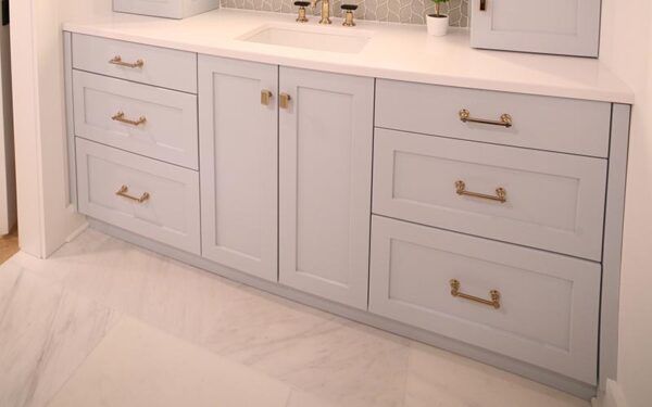 Light gray bathroom vanity with drawers and gold handles on a white marble floor