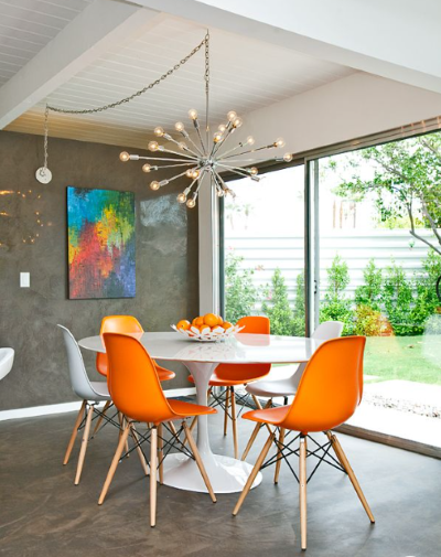 Modern dining room with orange chairs, round white table, colorful wall art, and garden view
