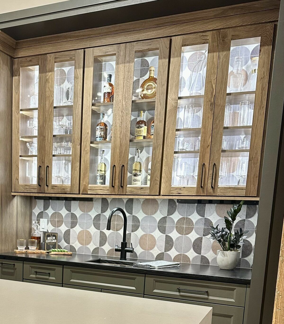 Wooden kitchen cabinets with glass doors, patterned tile backsplash, sink, and potted plants on a black countertop