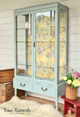 Mint green glass-front cabinet with floral wallpaper panels on a porch, beside flowers and a wooden crate