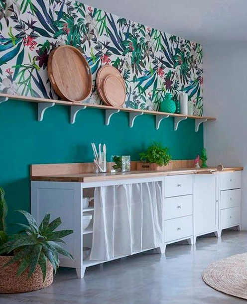 Modern kitchen with teal wall, floral wallpaper, wooden shelf, white cabinets, and wicker baskets.