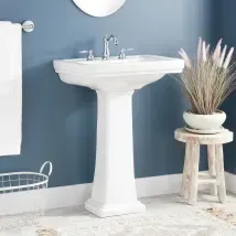 White pedestal sink in a blue bathroom with a towel rack, basket, and stool holding a plant.