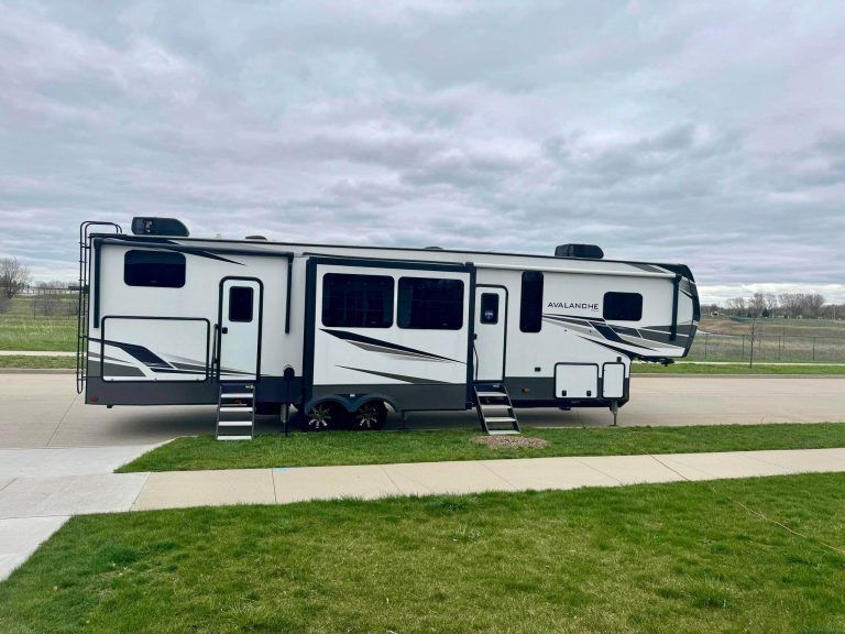White and gray fifth-wheel RV parked by a sidewalk under a cloudy sky
