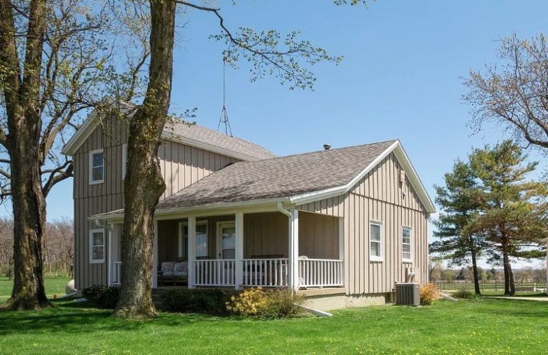 Small beige farmhouse with a porch and white railings, surrounded by green lawn and trees under a blue sky