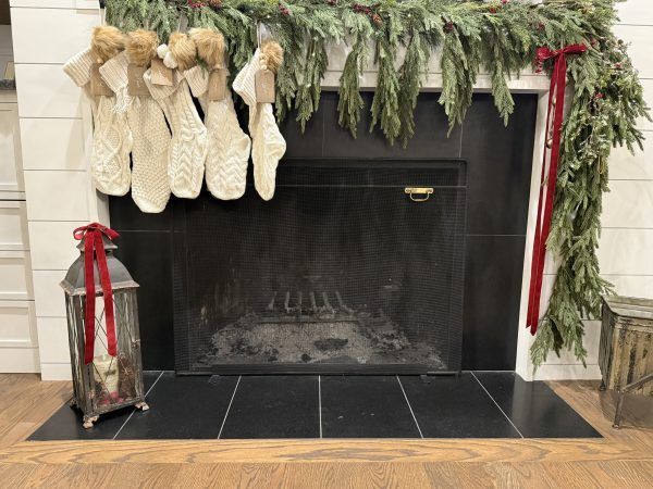 Christmas stockings and greenery draped over a black fireplace mantel with a lantern beside it
