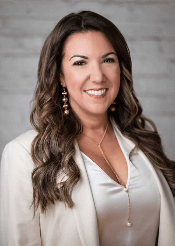 Smiling woman with long wavy hair in a white blouse and beige blazer against a gray wood backdrop