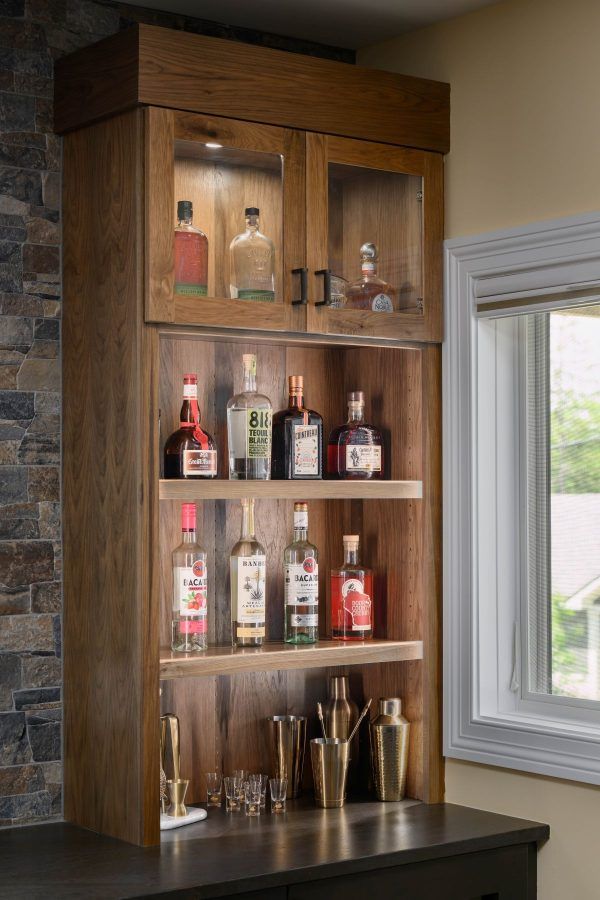Wooden home bar cabinet with shelves of liquor bottles and glassware by a window
