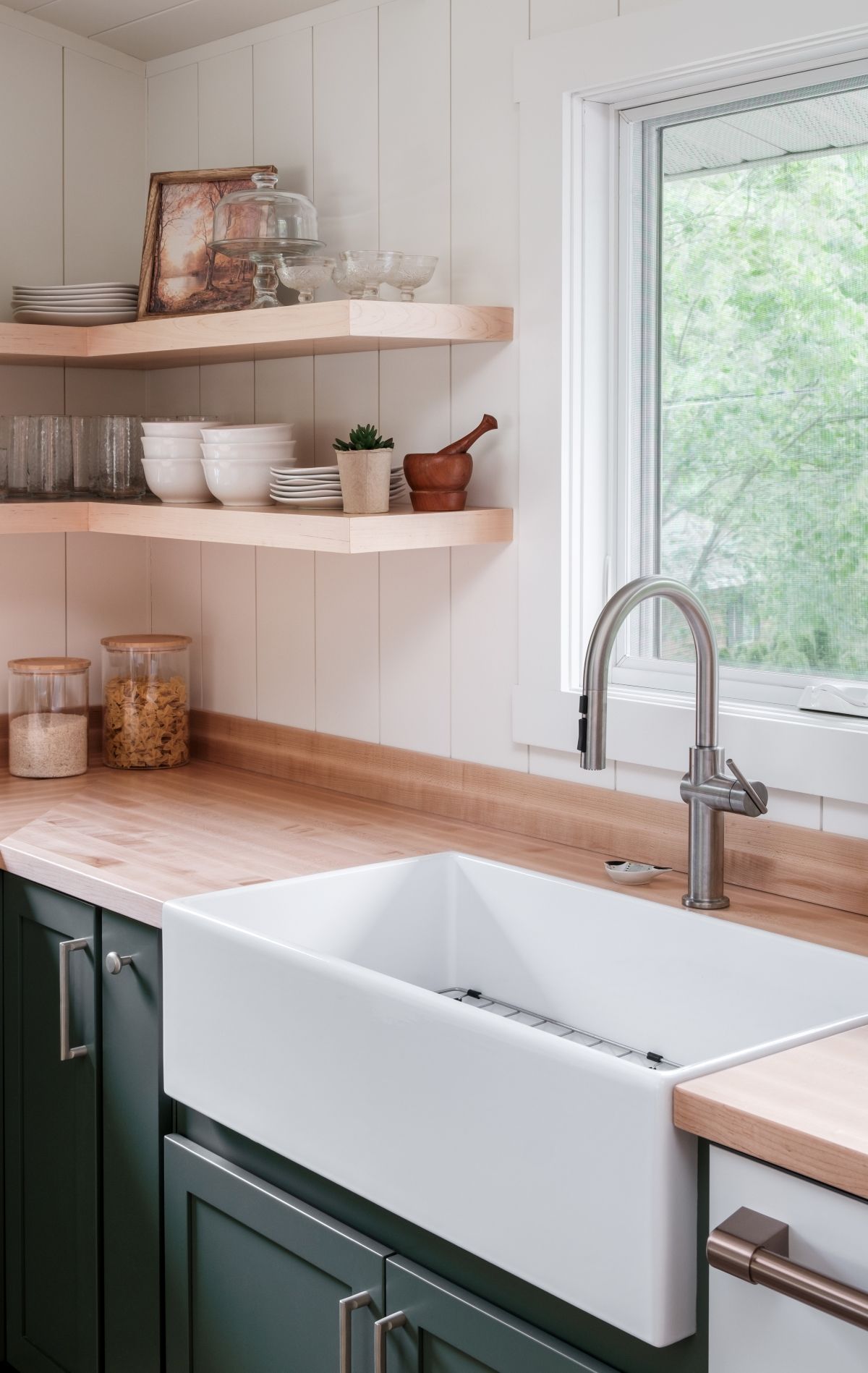 Modern kitchen sink with white apron-front basin, dark green cabinets, wood countertops, and open shelves by a window