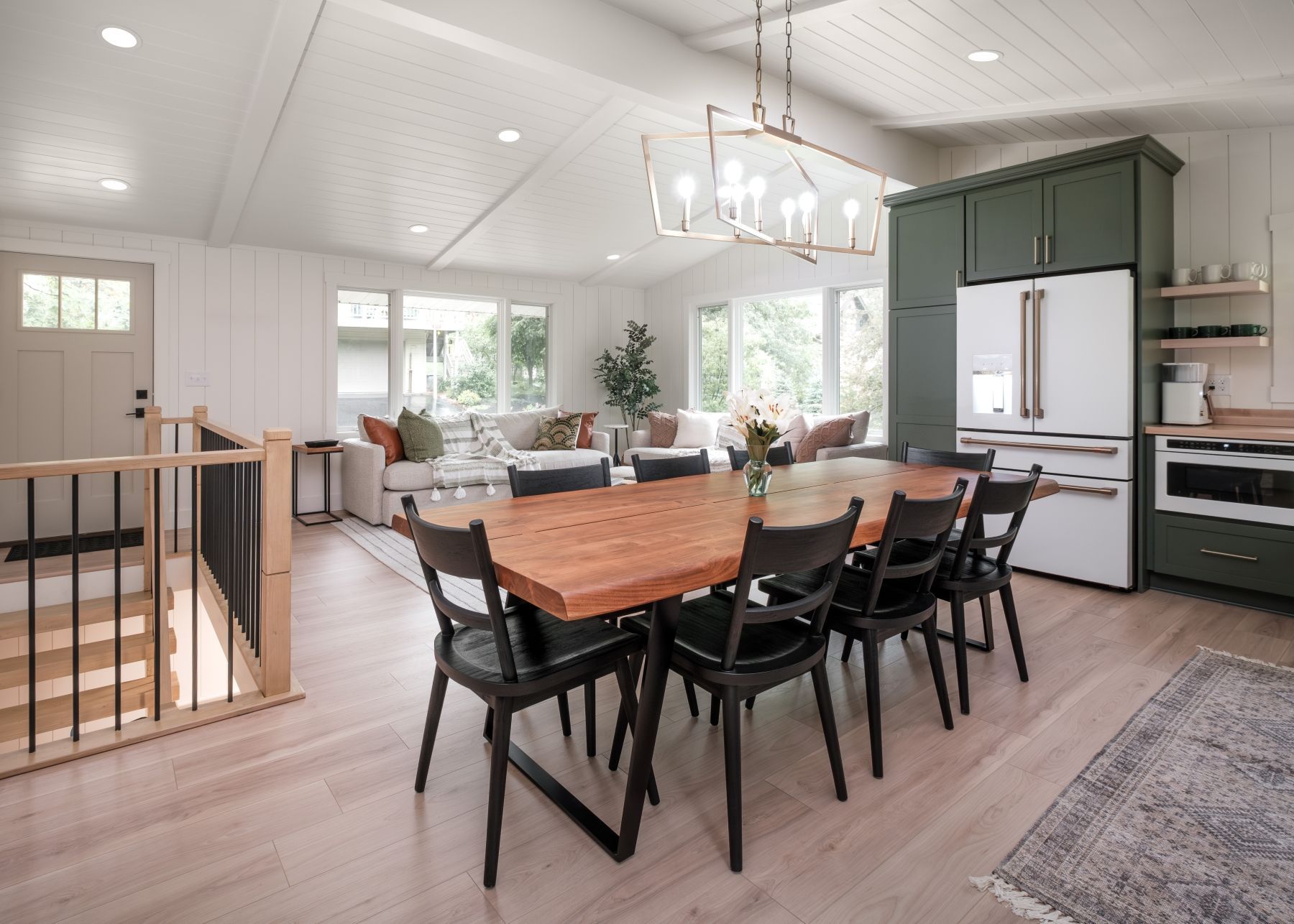 Modern open-plan kitchen and dining area with wooden table, black chairs, green cabinets, and bright windows