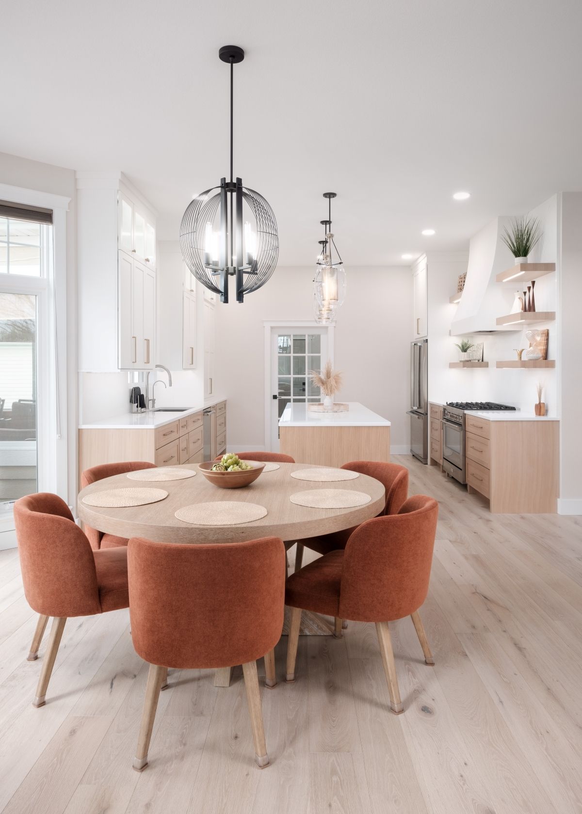 Bright modern kitchen and dining area with round table, rust chairs, white cabinets, and pendant lights