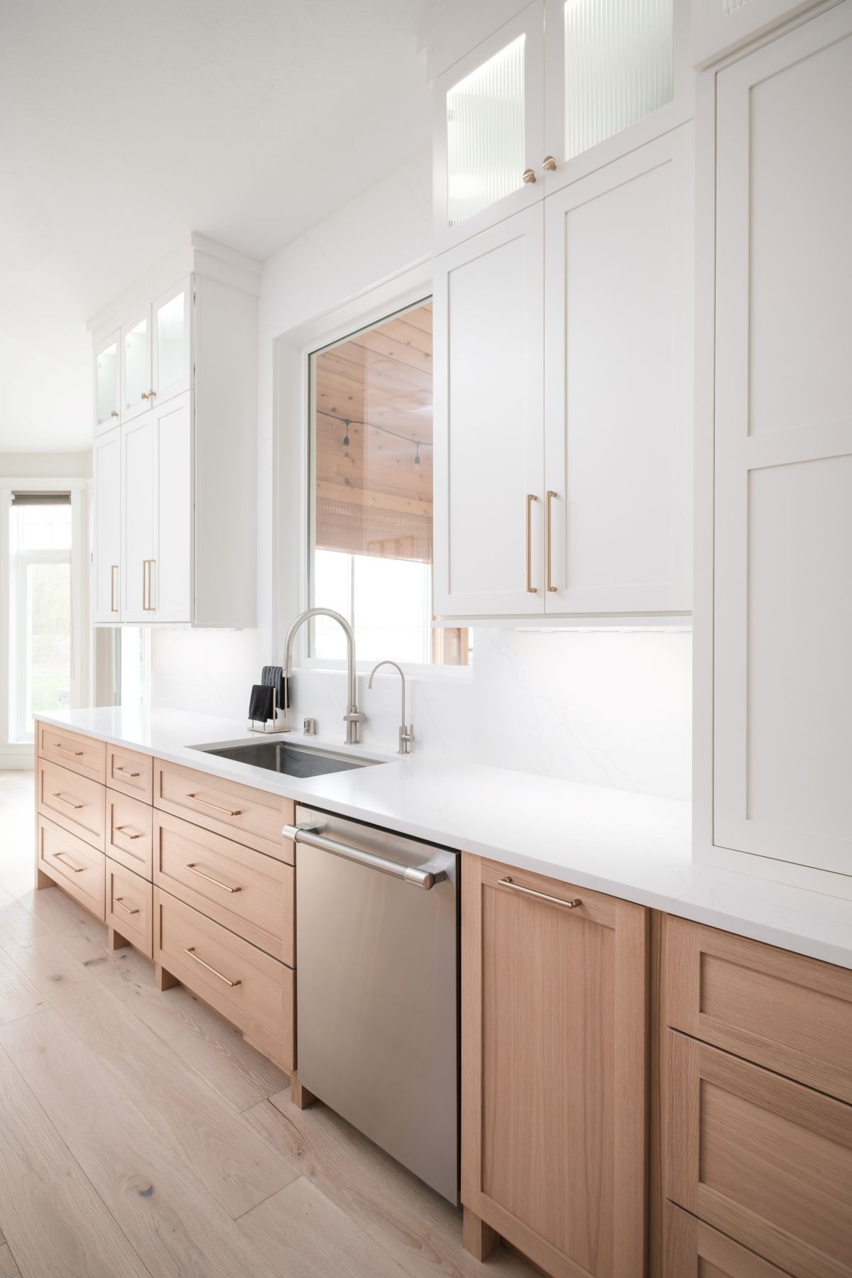 Bright modern kitchen with white cabinets, wood lower drawers, stainless dishwasher, and sink under a window.
