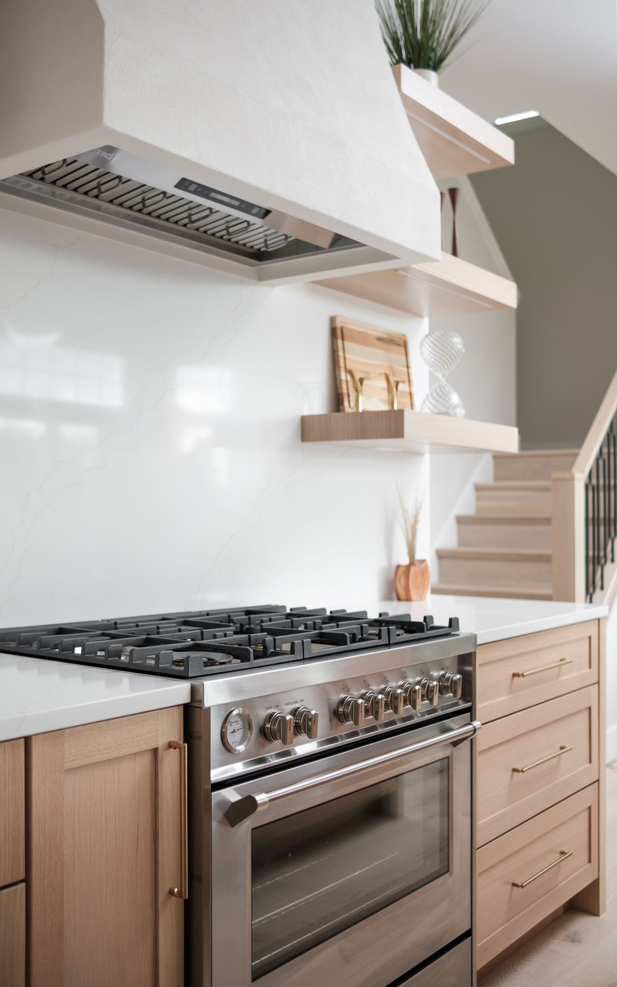 Modern kitchen with stainless steel range, light wood cabinets, white walls, and floating shelves