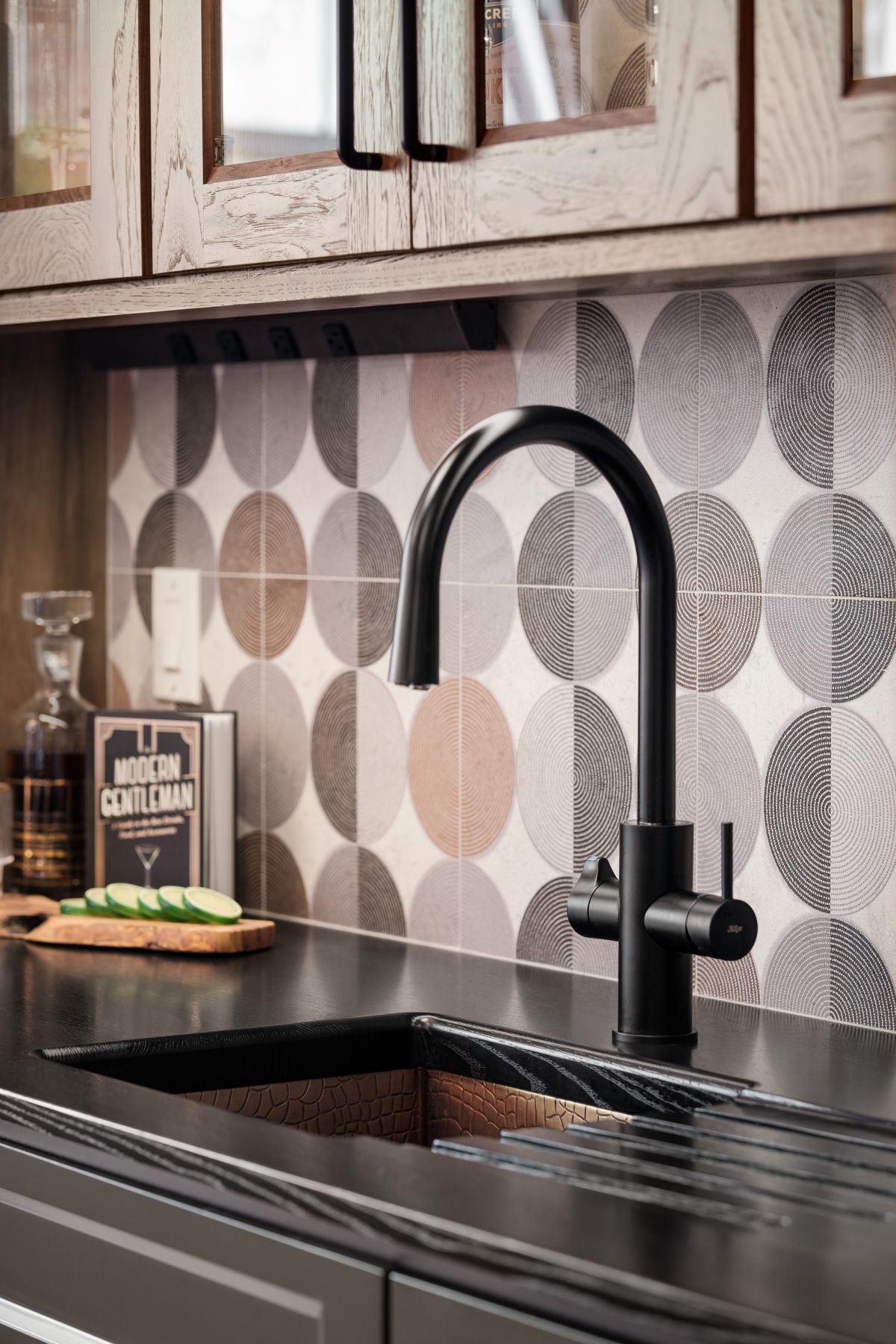 Modern kitchen sink with tall black faucet, patterned tile backsplash, and wooden cabinets.