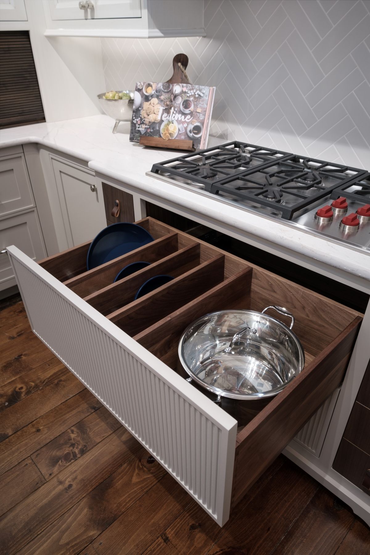 Open drawer with wood utensil organizer and metal mixing bowl in a white kitchen beside a gas stove