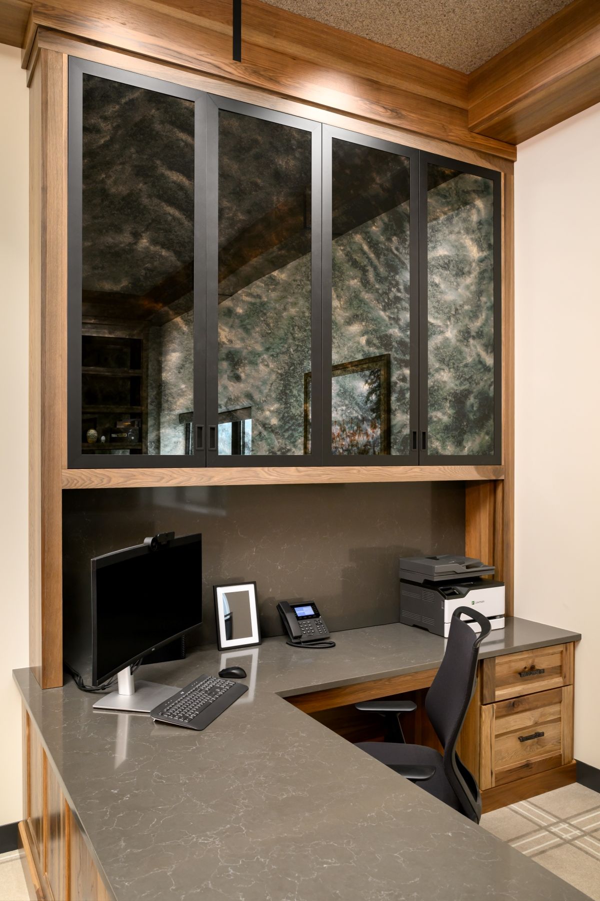 Small office desk with computer, monitor, printer, and chair beneath a wall of large windows.