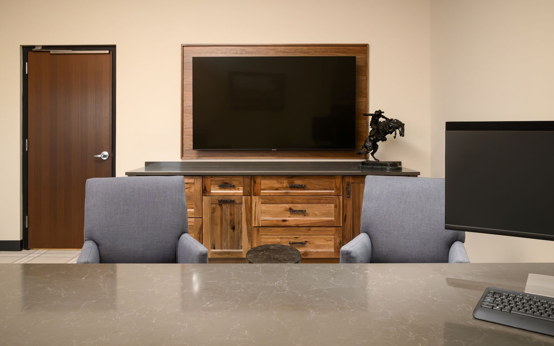 Office with a desk, two gray chairs, a TV on a cabinet, and a closed wooden door.
