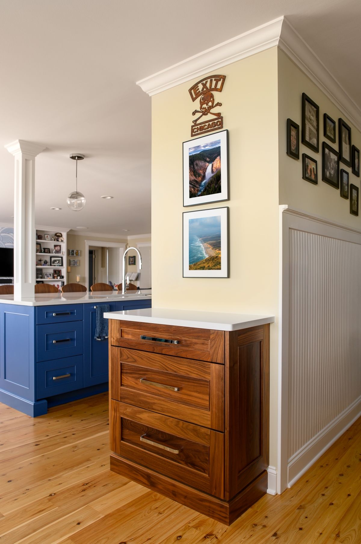 Hallway corner with wooden dresser, framed photos, and a blue kitchen visible nearby