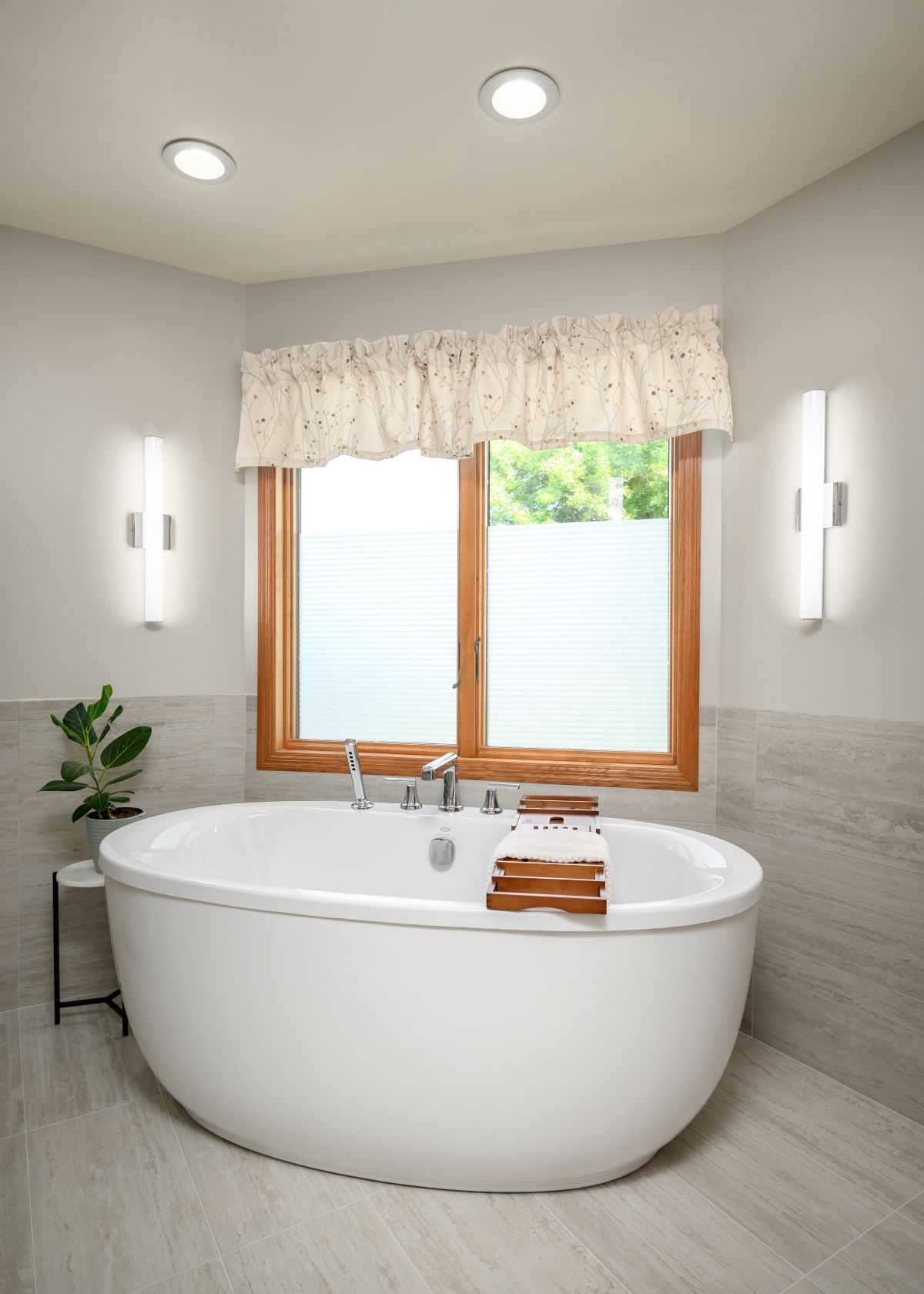 Bright white bathroom with a freestanding tub, wood-framed window, and wall sconces under a ruffled curtain.