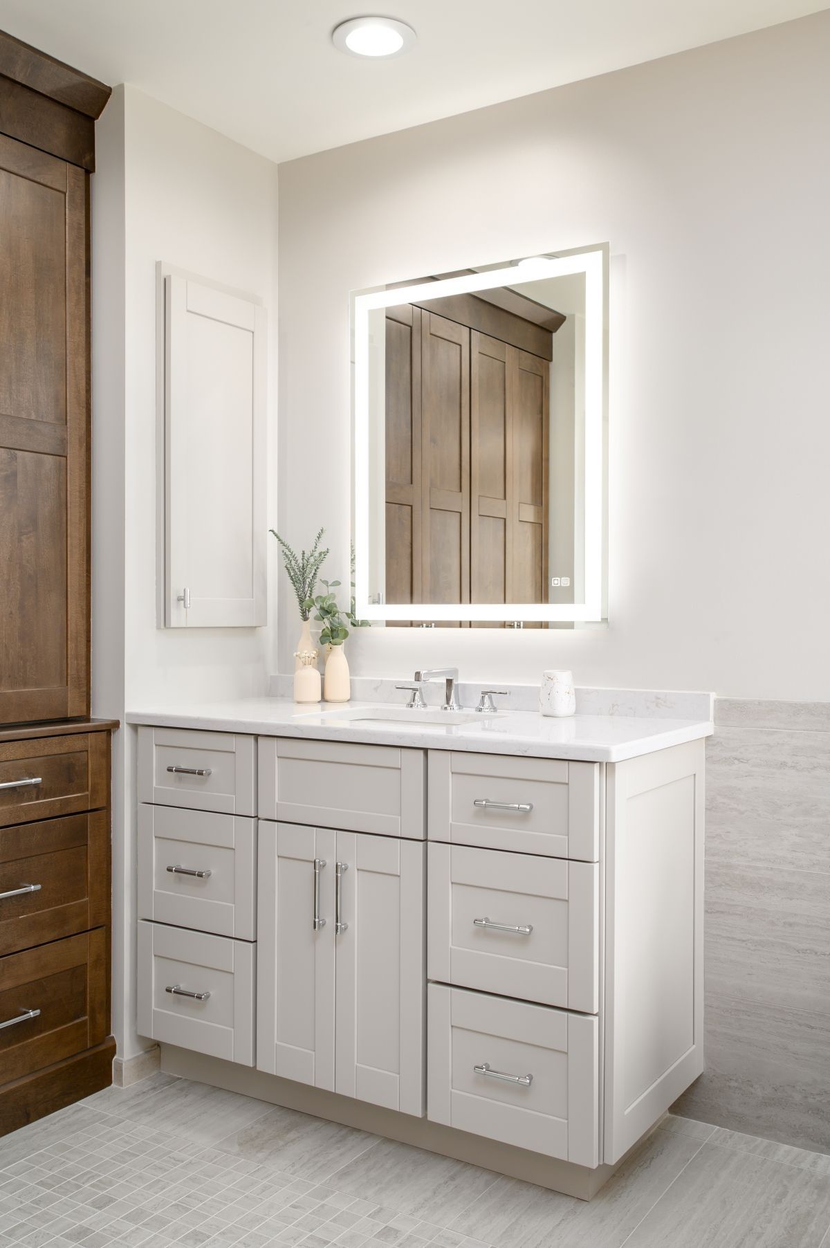 Modern bathroom vanity with white countertop, lit mirror, and gray-beige tiled wall.
