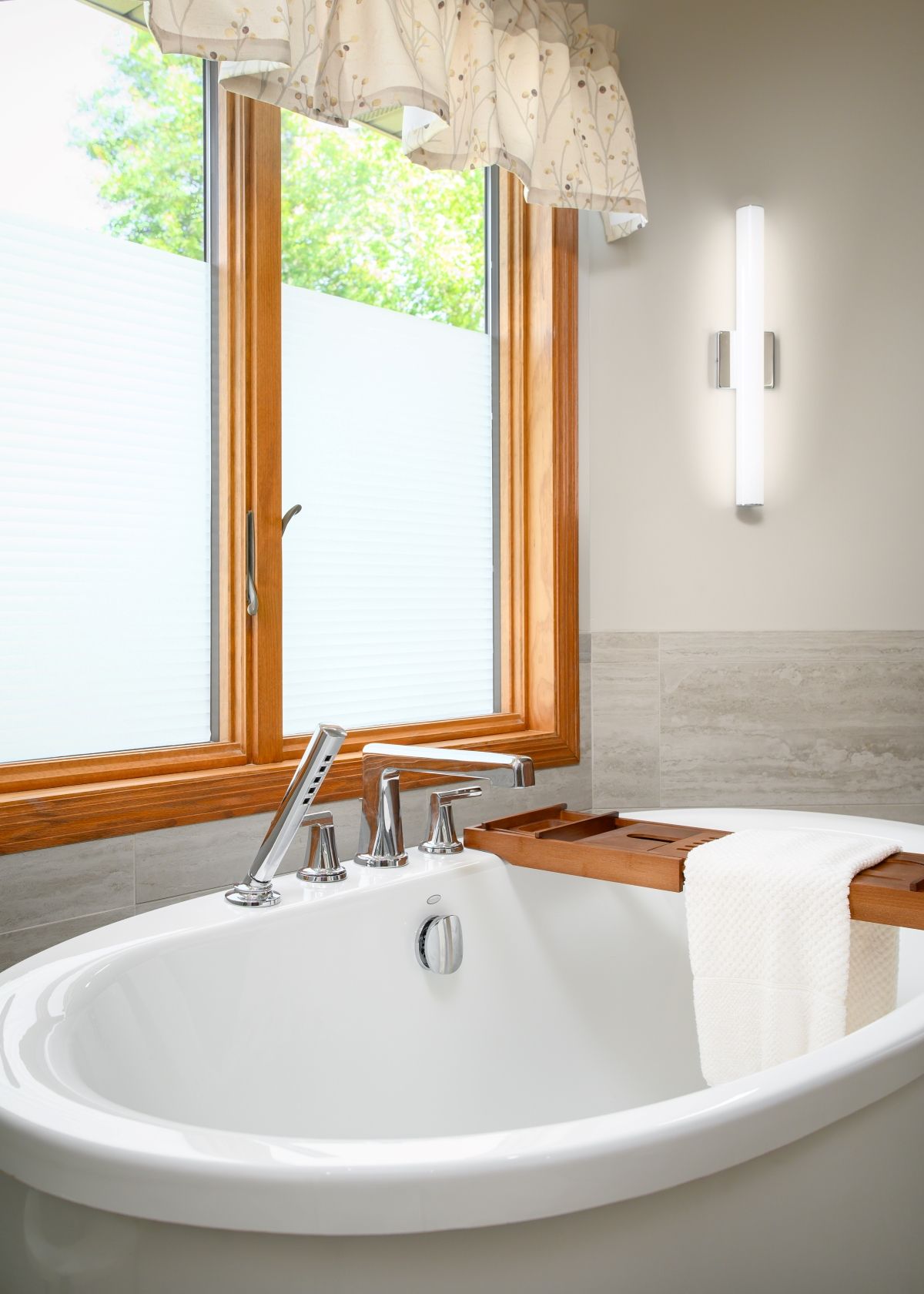 Bright bathroom with a white soaking tub, wood-framed window, and wall sconce beside a sink.