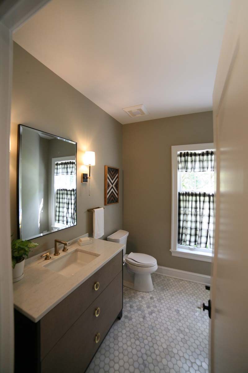 Small bathroom with gray vanity, wall mirror, toilet, and window with patterned curtains.