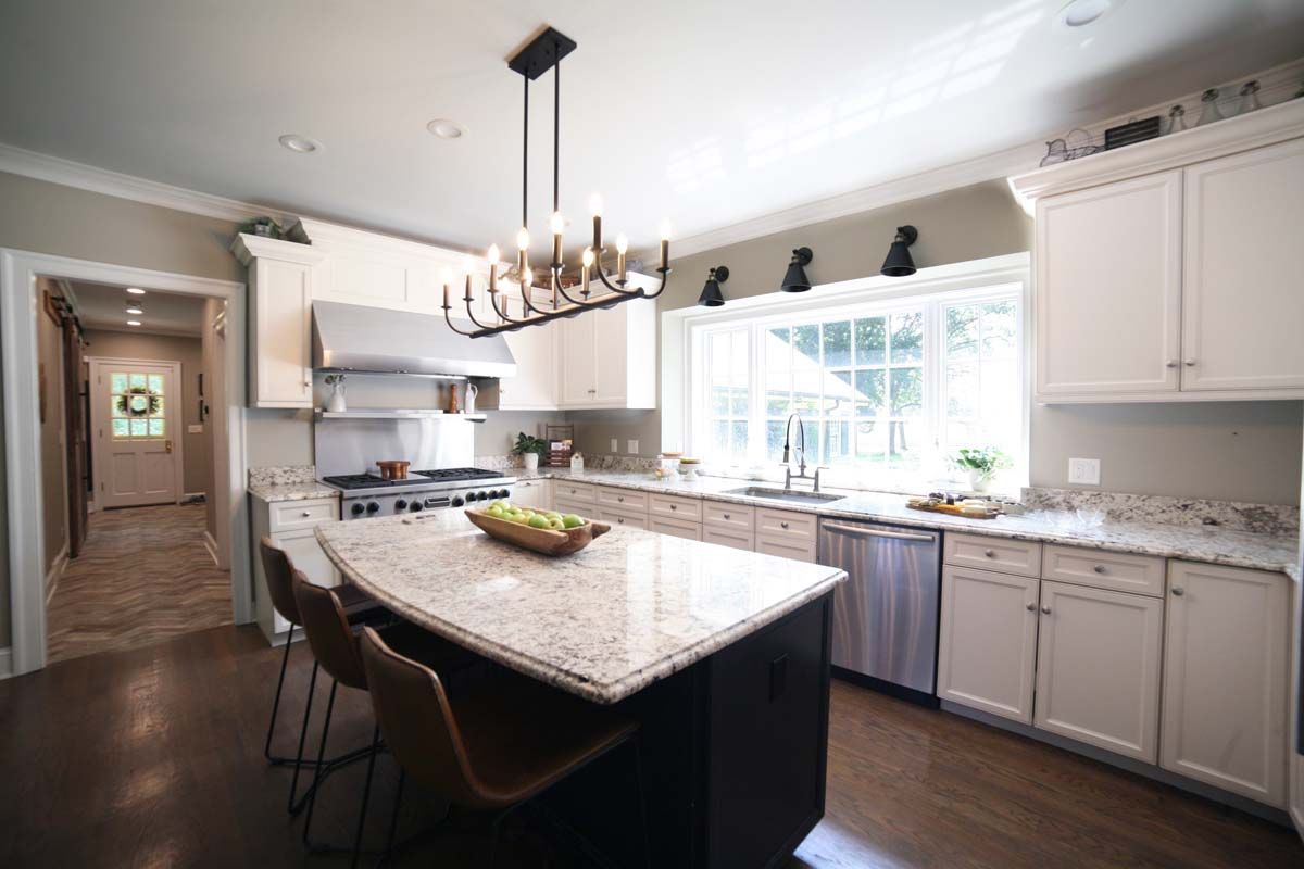 Modern kitchen with large island, white cabinets, granite counters, and a chandelier by a bright window.