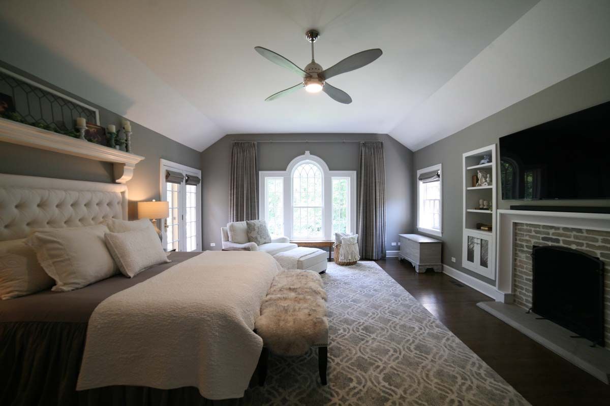 Spacious gray bedroom with tufted bed, fireplace, TV, and large arched window with curtains