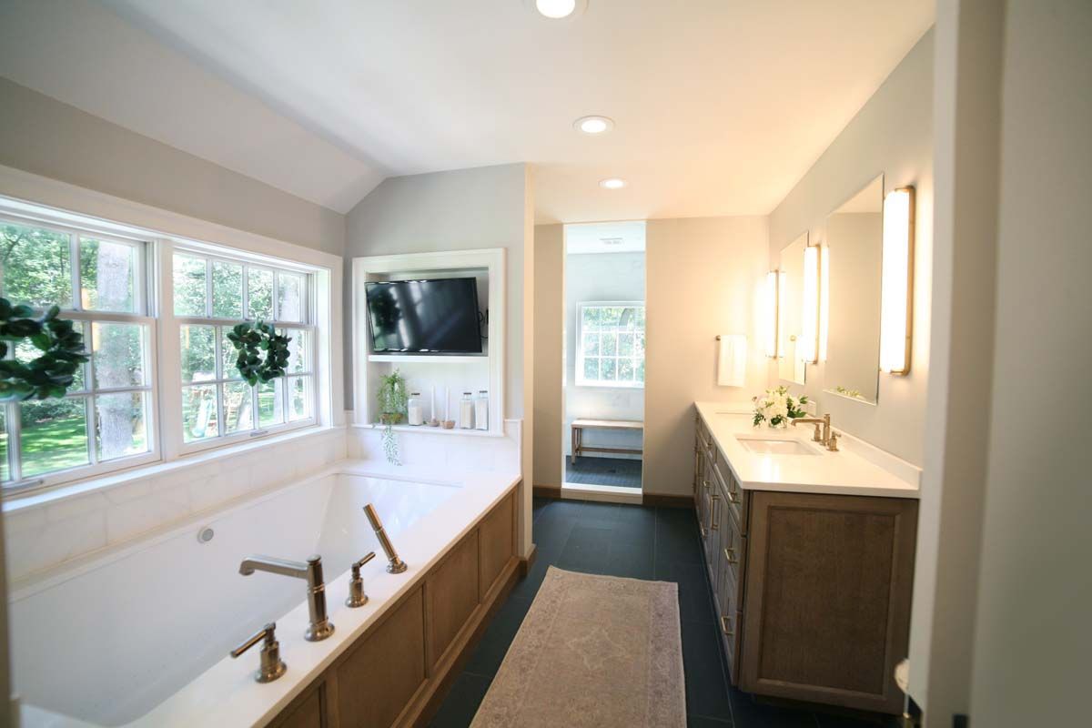 Bright bathroom with long vanity, large window, dual sinks, mirrors, and a freestanding bathtub by the wall