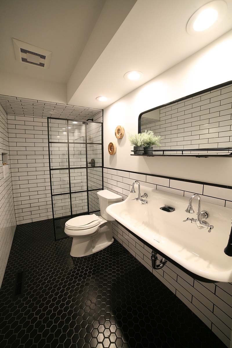 Modern white-tiled bathroom with double sinks, black hex floor, toilet, and glass shower stall