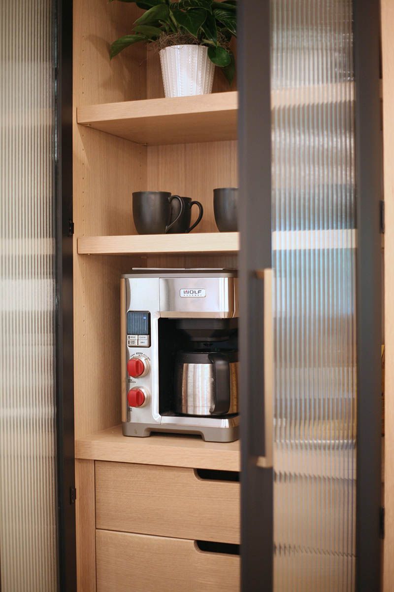 Coffee maker on a shelf in a kitchen nook, with a plant and mug above and drawers below.