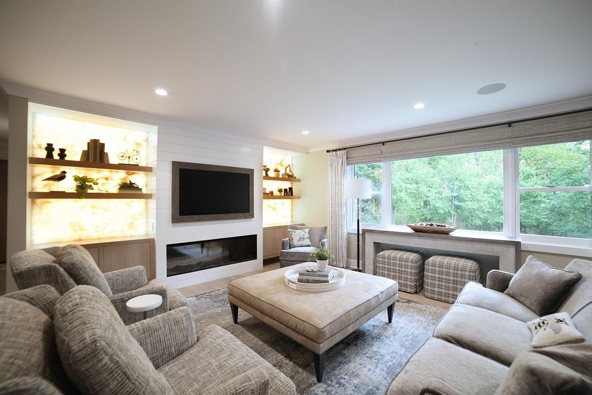 Bright modern living room with gray sofas, wall-mounted TV, fireplace, and large windows overlooking greenery