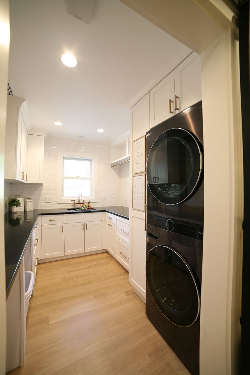 Bright galley kitchen with white cabinets, black countertops, and stacked black washer-dryer units