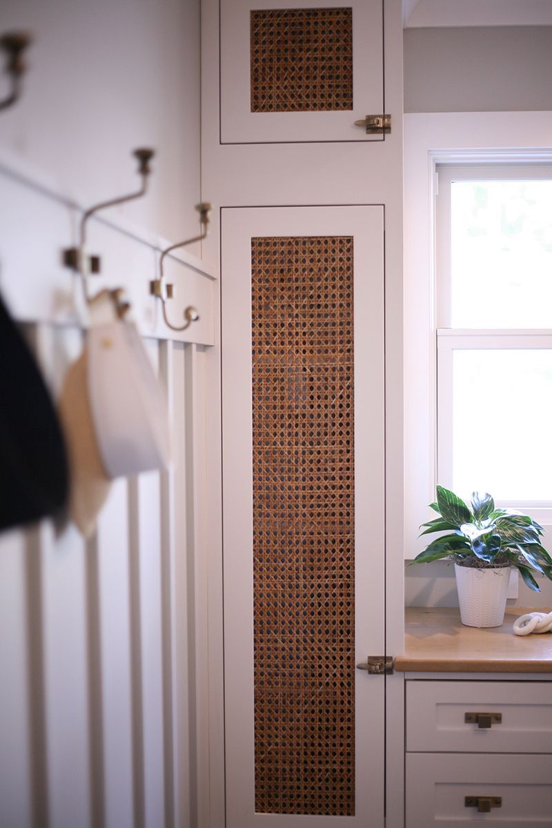 White room with wicker vent covers, coat rack, and a dresser with a potted plant by a window.