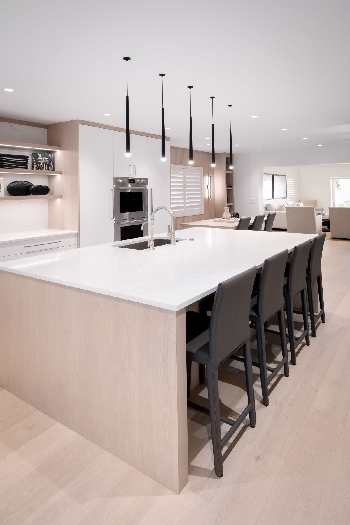 Modern open kitchen with large white island, black stools, pendant lights, and light wood cabinetry.