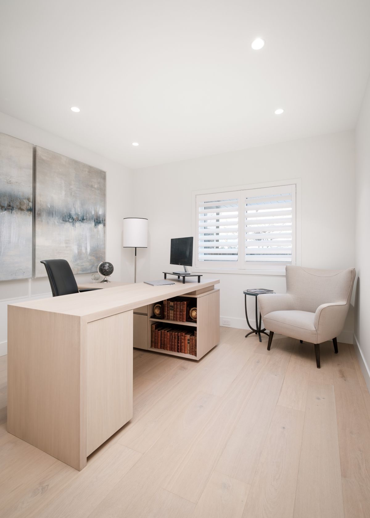 Modern home office with a light wood desk, beige chair, and bright white walls and shutters.