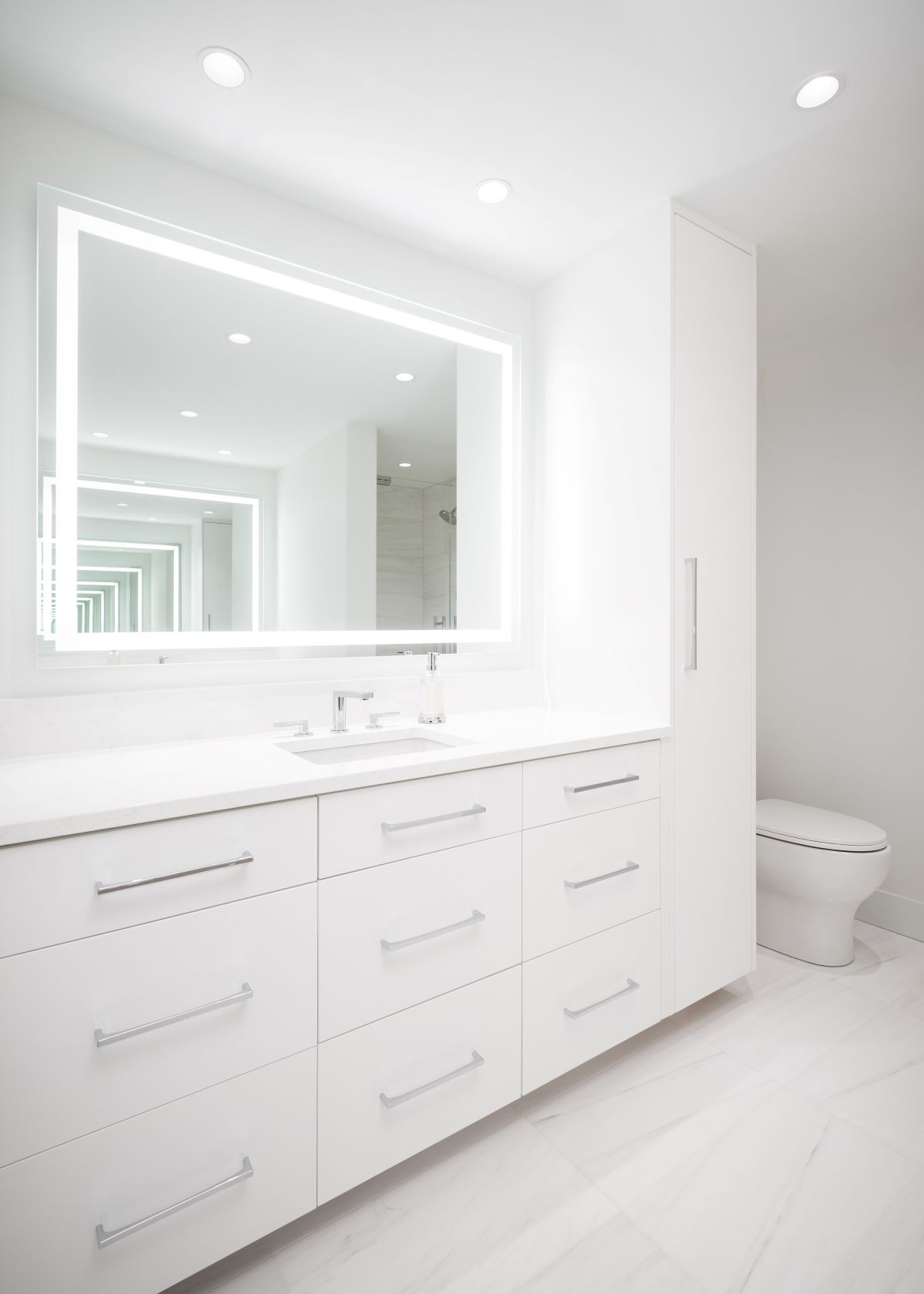 Bright white bathroom vanity with illuminated mirror, sink, drawers, and toilet in a minimalist room