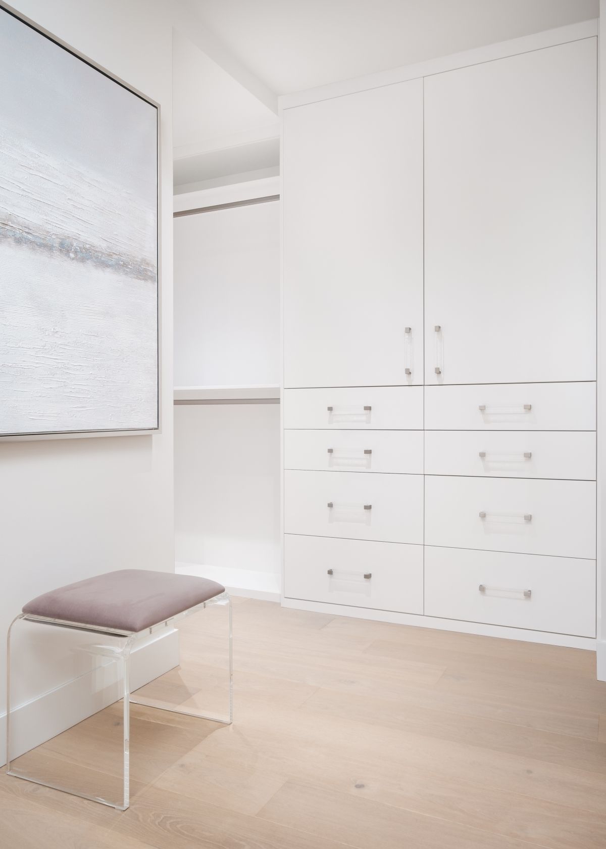 Minimal white bedroom with built-in closets, drawers, and a small cushioned stool by the wall.
