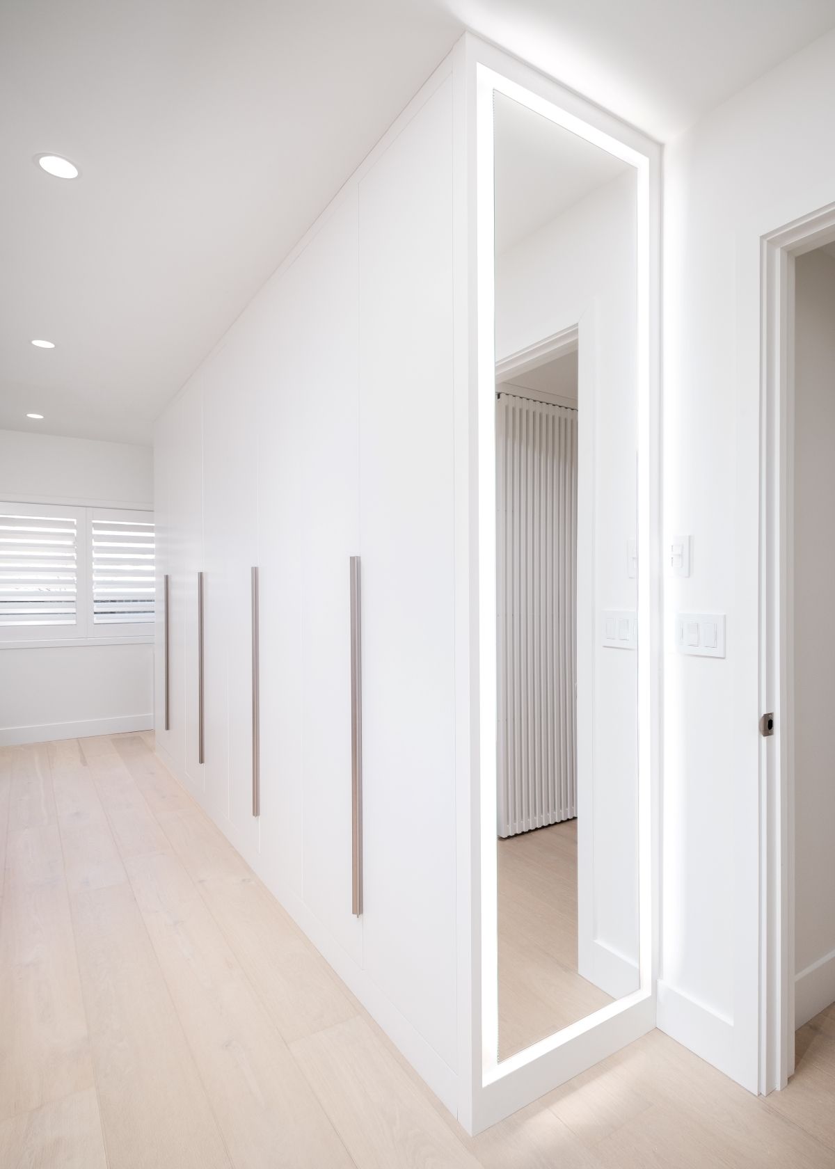 Bright white hallway with recessed lights, wall trim, and a full-length mirror beside a doorway.